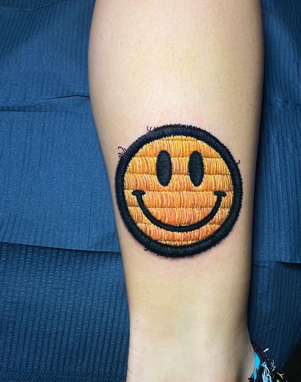 Embroidered patch-style yellow smiley face tattoo on calf