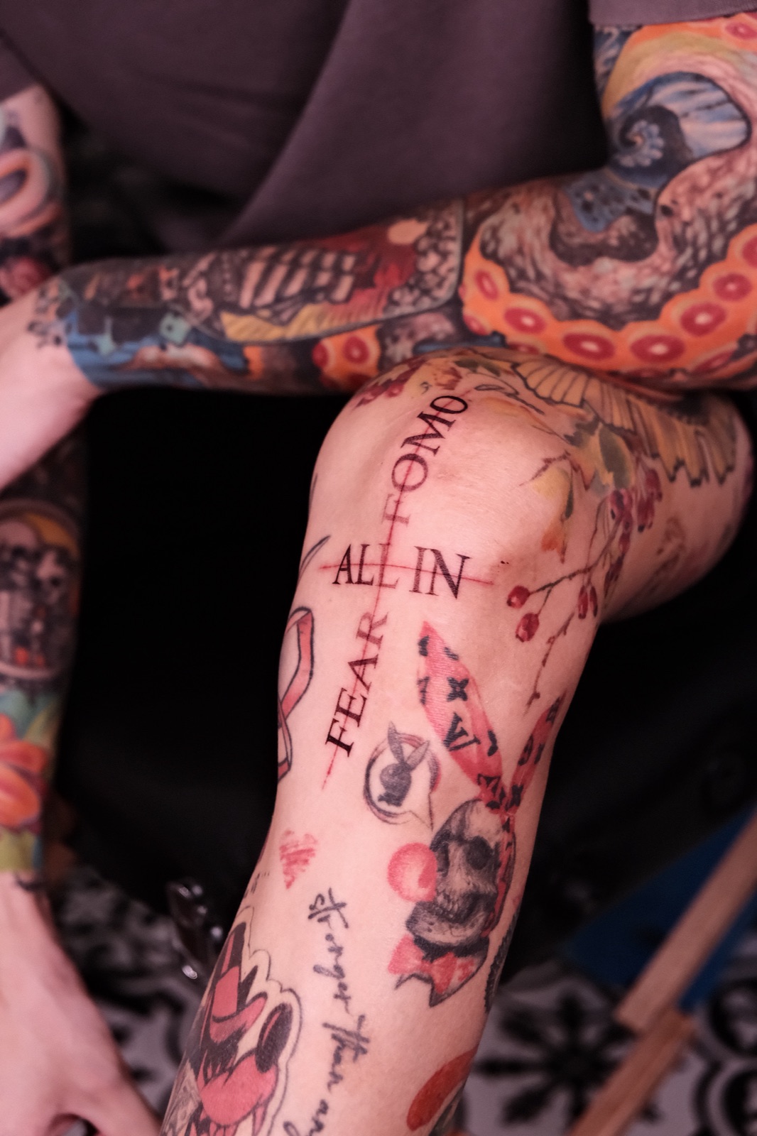 Lettering tattoo reading ALL IN and FEAR FOMO among other pieces on the forearm