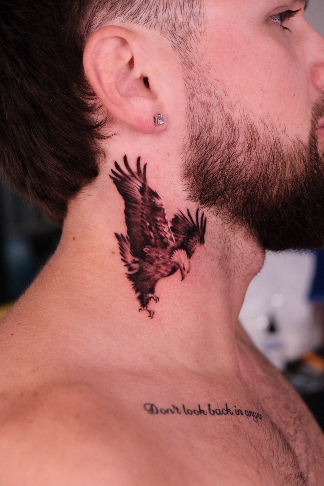 Black and grey realistic tattoo of a landing eagle on the side of the neck, with cursive lettering below on the collarbone