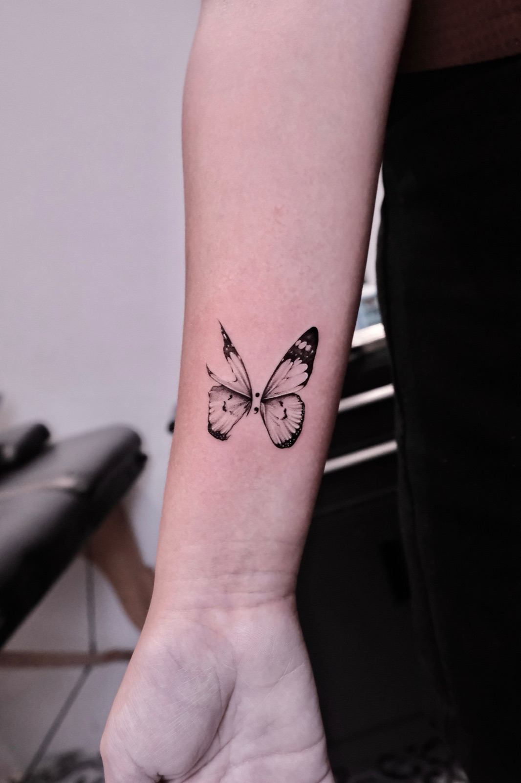 Black-and-grey butterfly tattoo with a semicolon symbol between the wings on the wrist