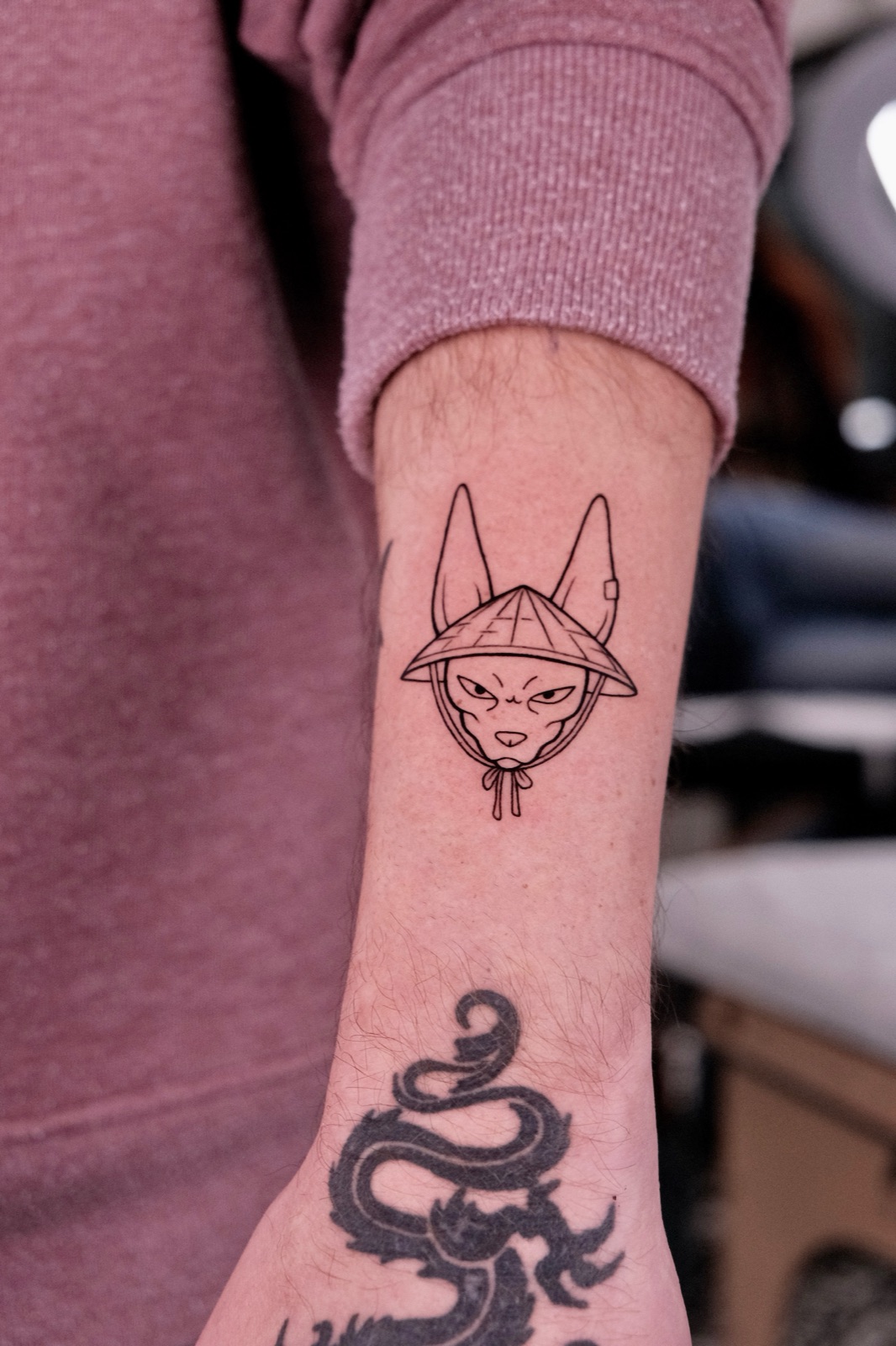 Fine-line tattoo of a cat wearing a Vietnamese conical hat on the forearm