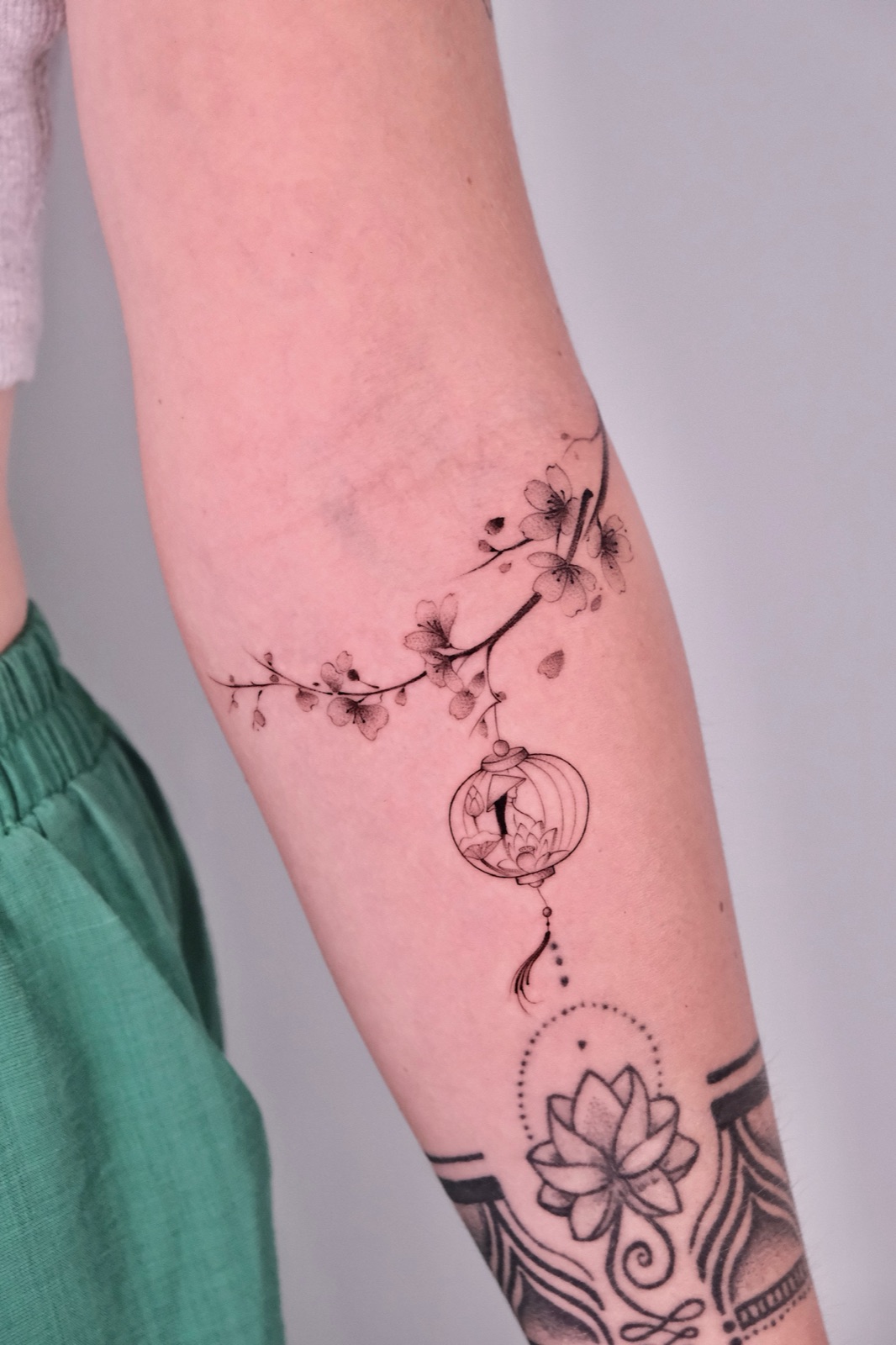 Fine-line cherry blossom branch tattoo draping down to a small lantern with a lotus inside, on the forearm