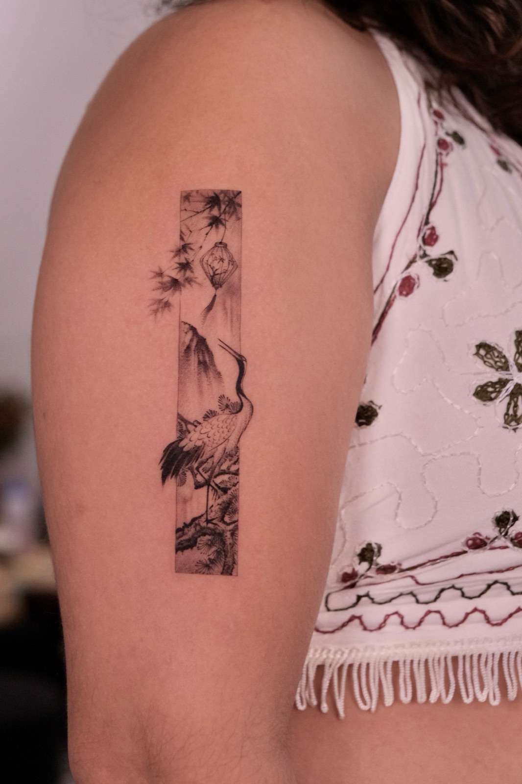 Black-and-grey tattoo panel of a crane beneath a hanging lantern and mountain landscape on the upper arm