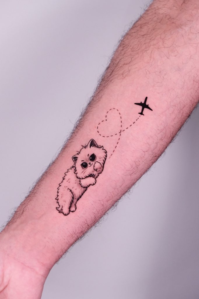 Tattoo of a fluffy cat with a heart-shaped flight path and airplane on forearm