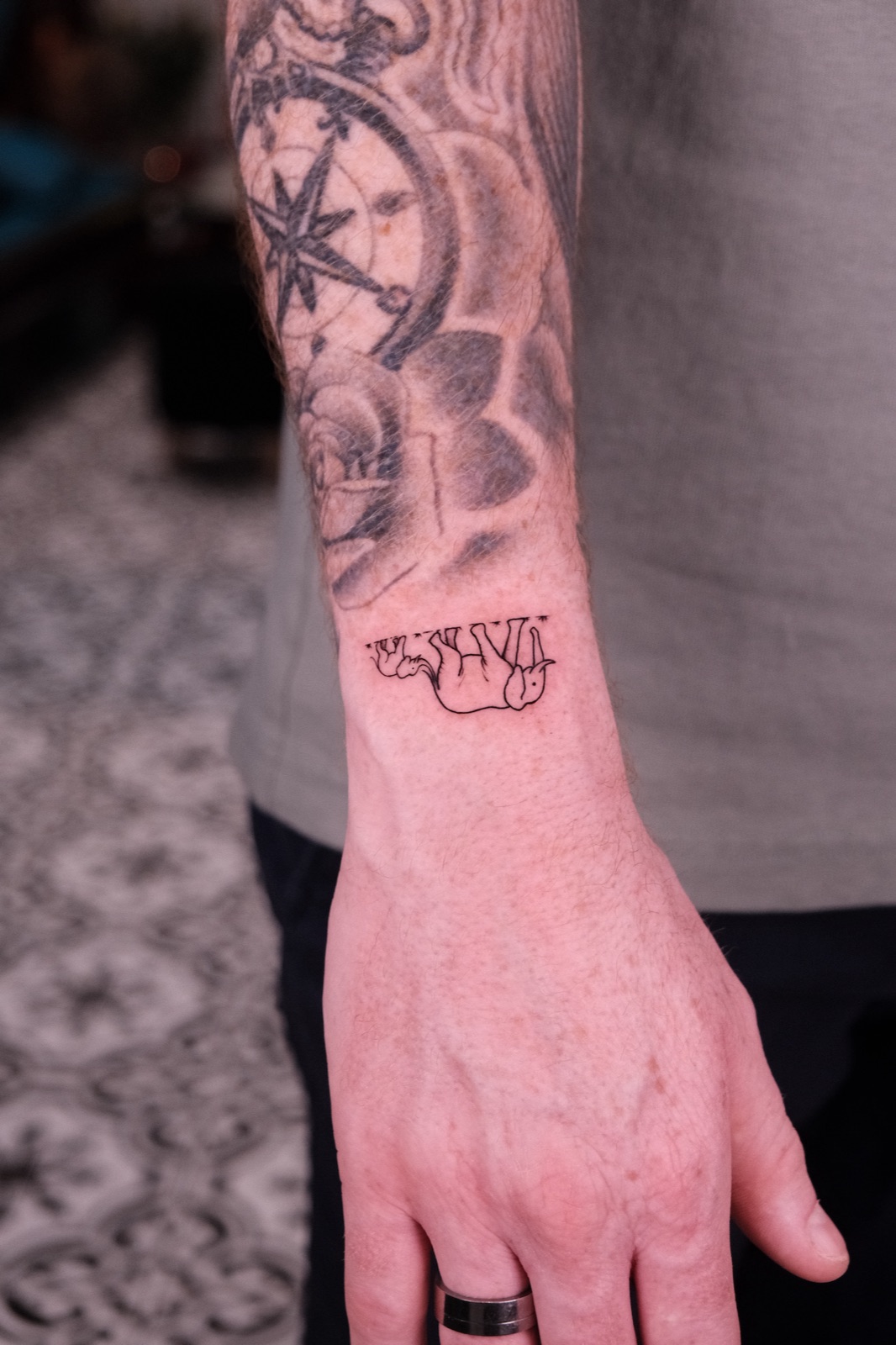Small fine-line tattoo of elephants hanging upside down with roman numerals on the wrist near a compass rose tattoo