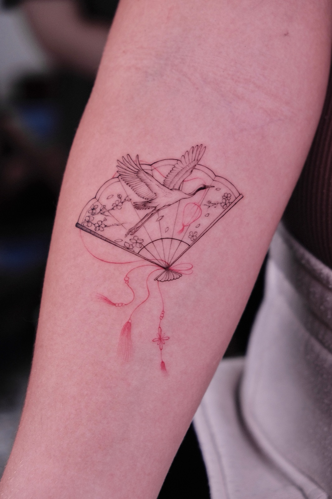 Fine-line fan tattoo featuring a flying crane, cherry blossoms and red tassels on the forearm