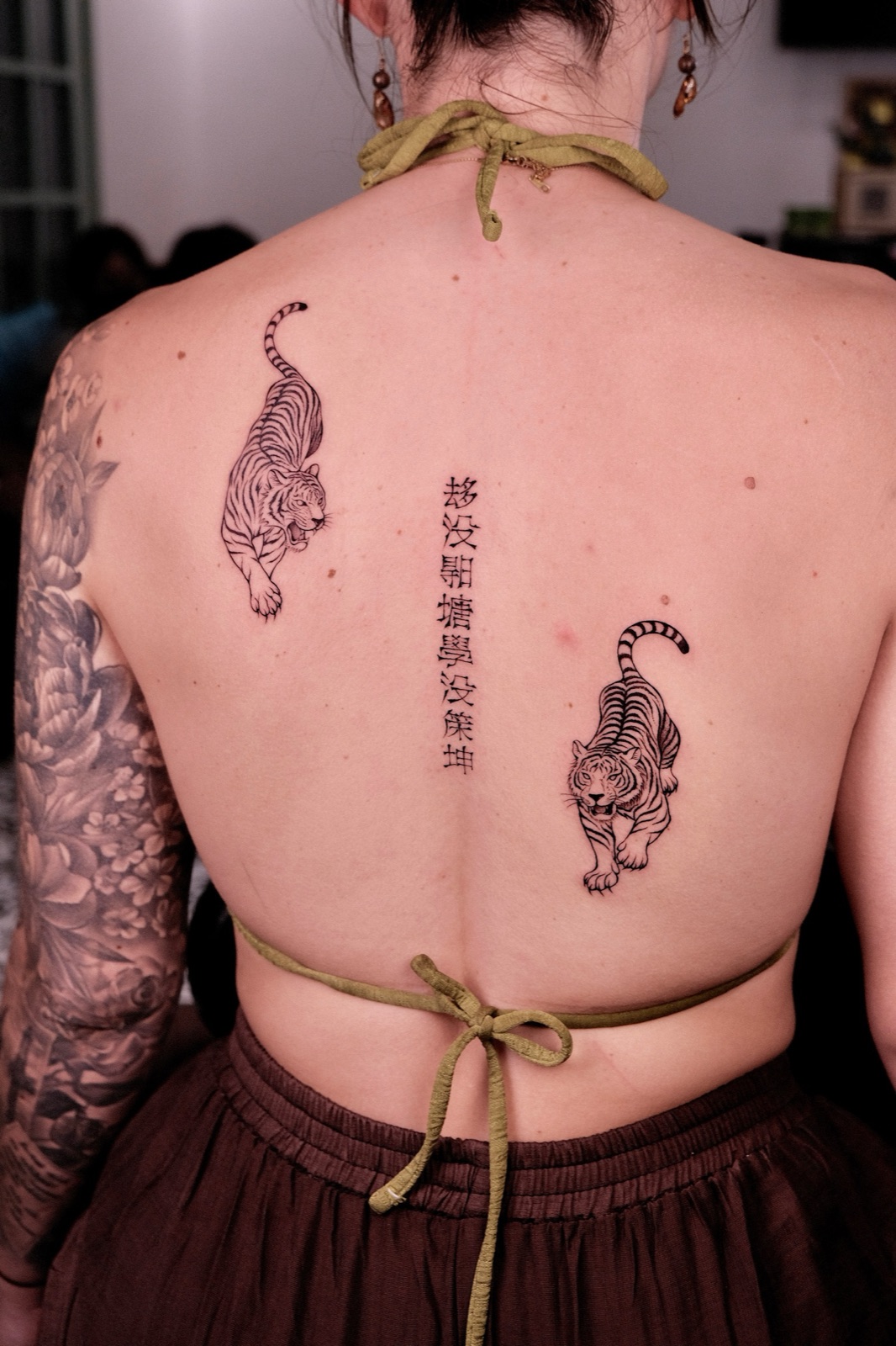 Matching fine-line tiger tattoos on the upper back separated by a vertical column of script
