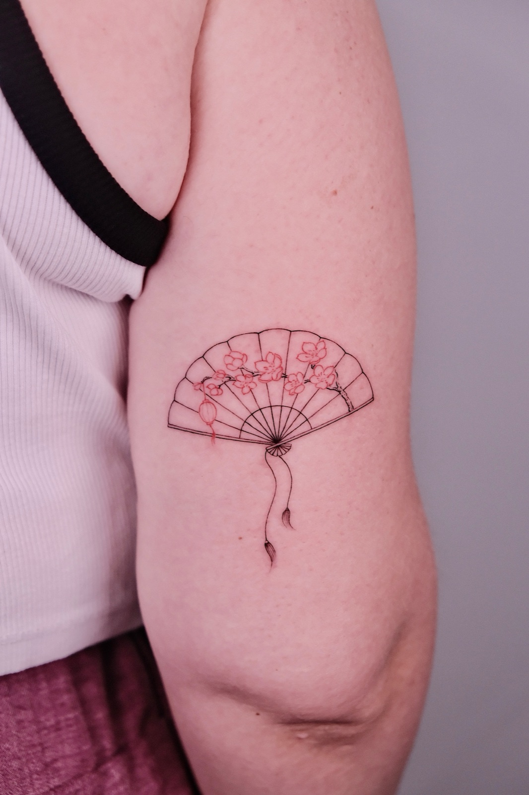 A fine-line folding fan tattoo with red cherry blossom outlines on the upper arm
