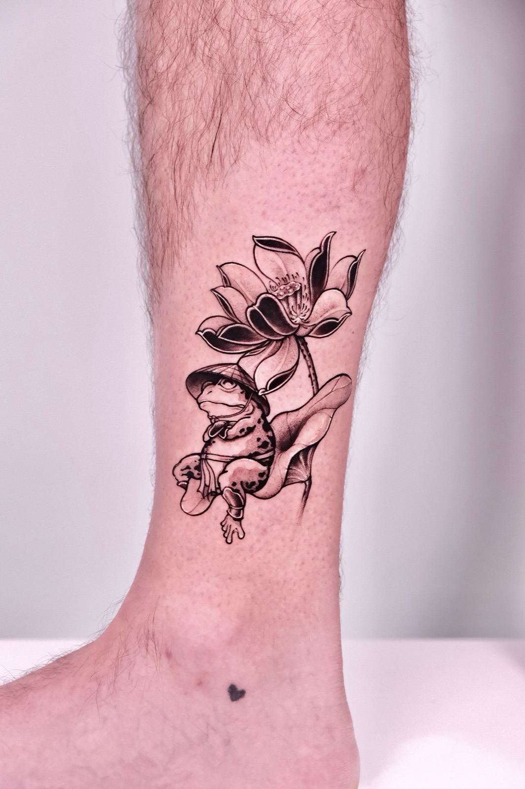 Tattoo of a frog wearing a conical hat sitting on a lotus leaf beside a lotus flower on the ankle