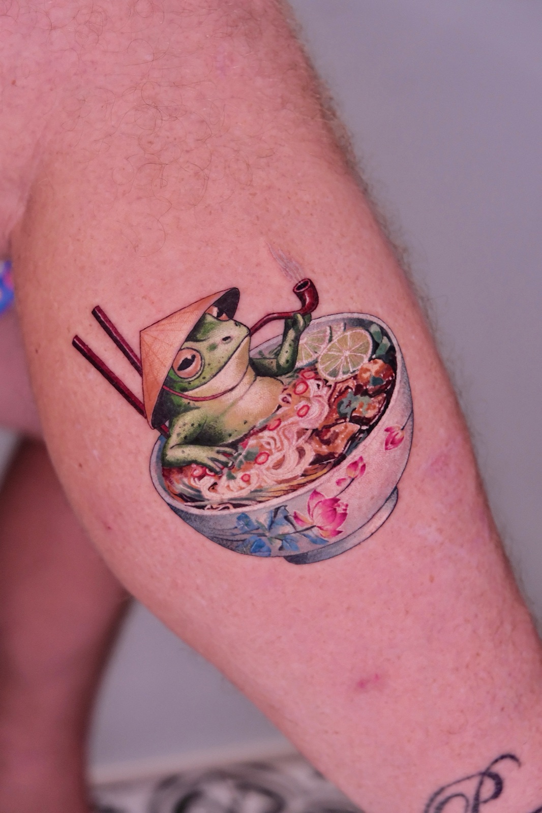 Colorful tattoo of a frog in a conical hat eating pho with chopsticks and a pipe on the leg