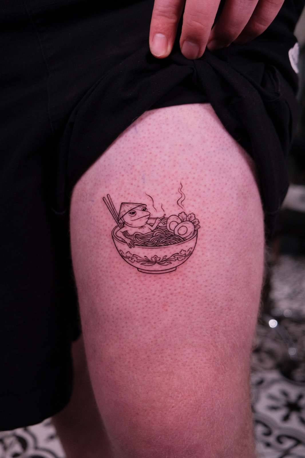 Fine-line tattoo of a frog in a conical hat eating pho on the thigh