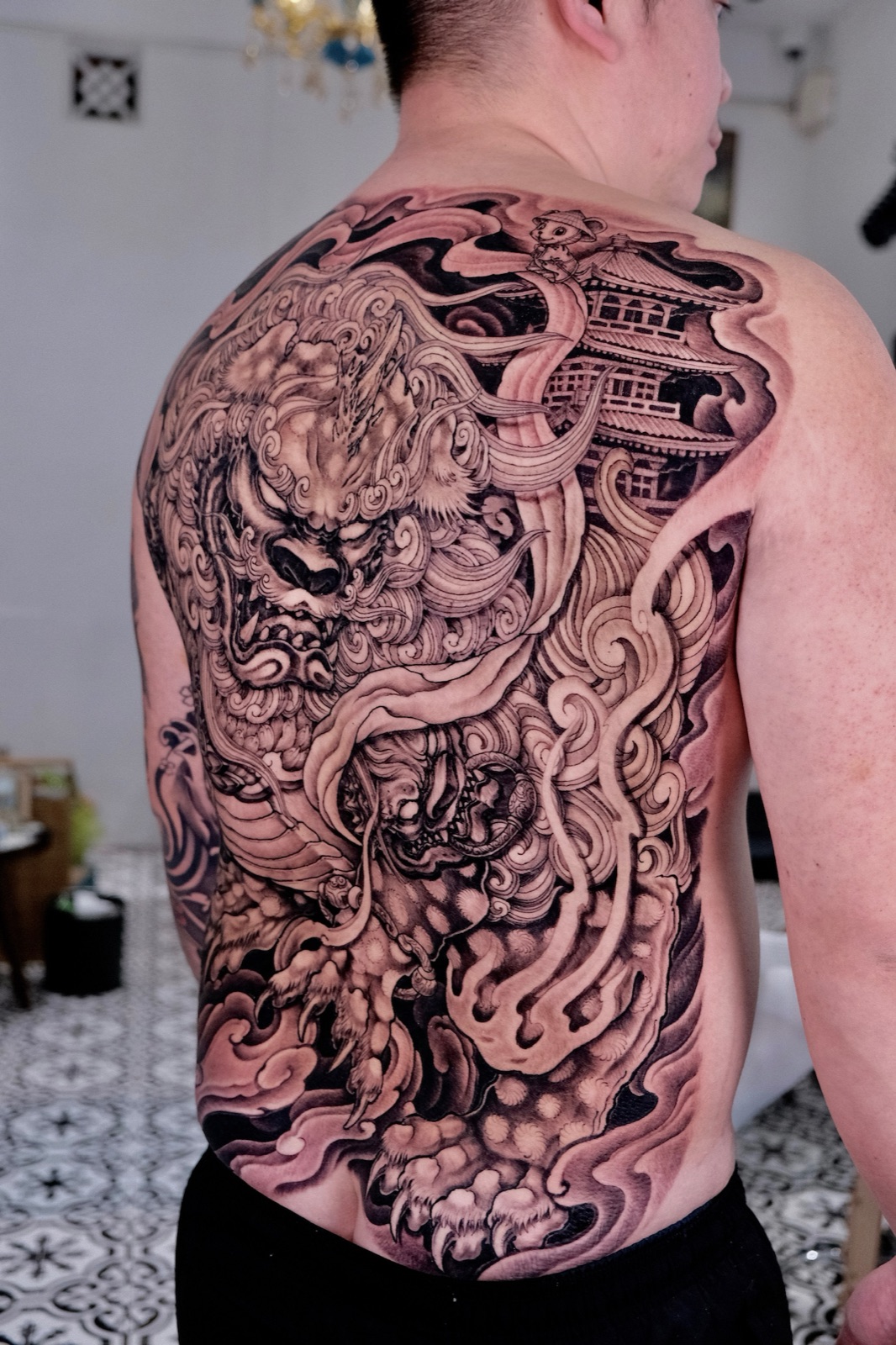 Black-and-grey Japanese-style backpiece featuring a guardian lion, clouds, and a pagoda