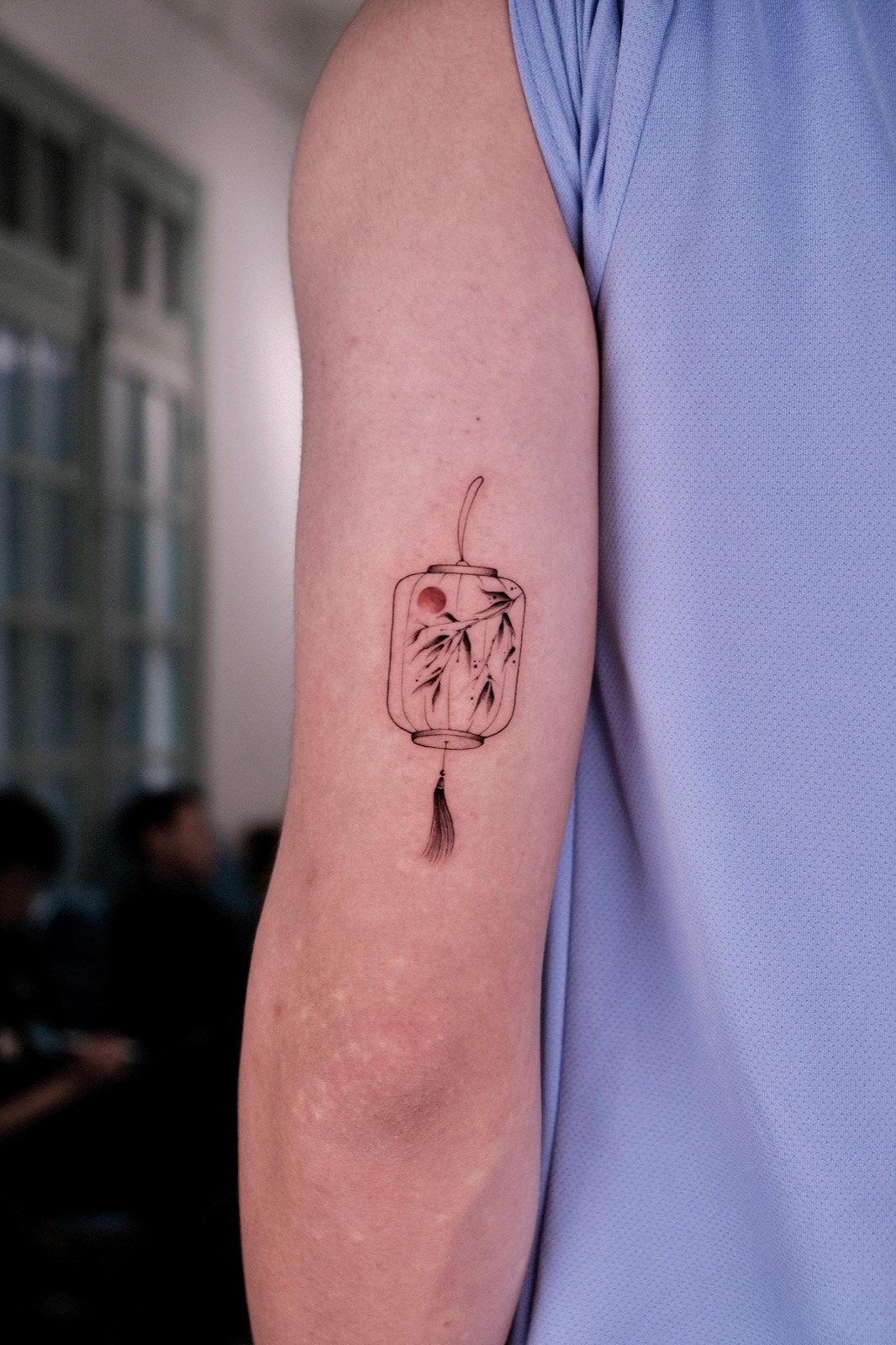 Fine-line hanging lantern tattoo with bamboo leaves on the upper arm