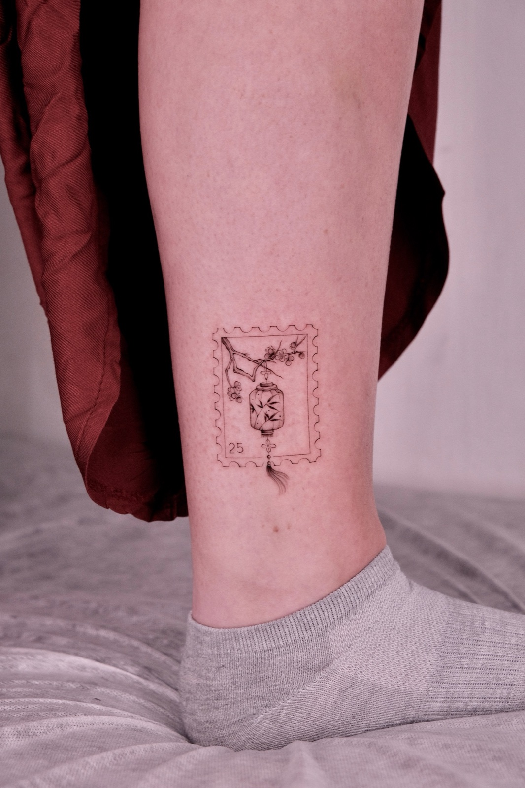 Fine-line postage stamp tattoo with a hanging lantern and cherry blossoms on the ankle