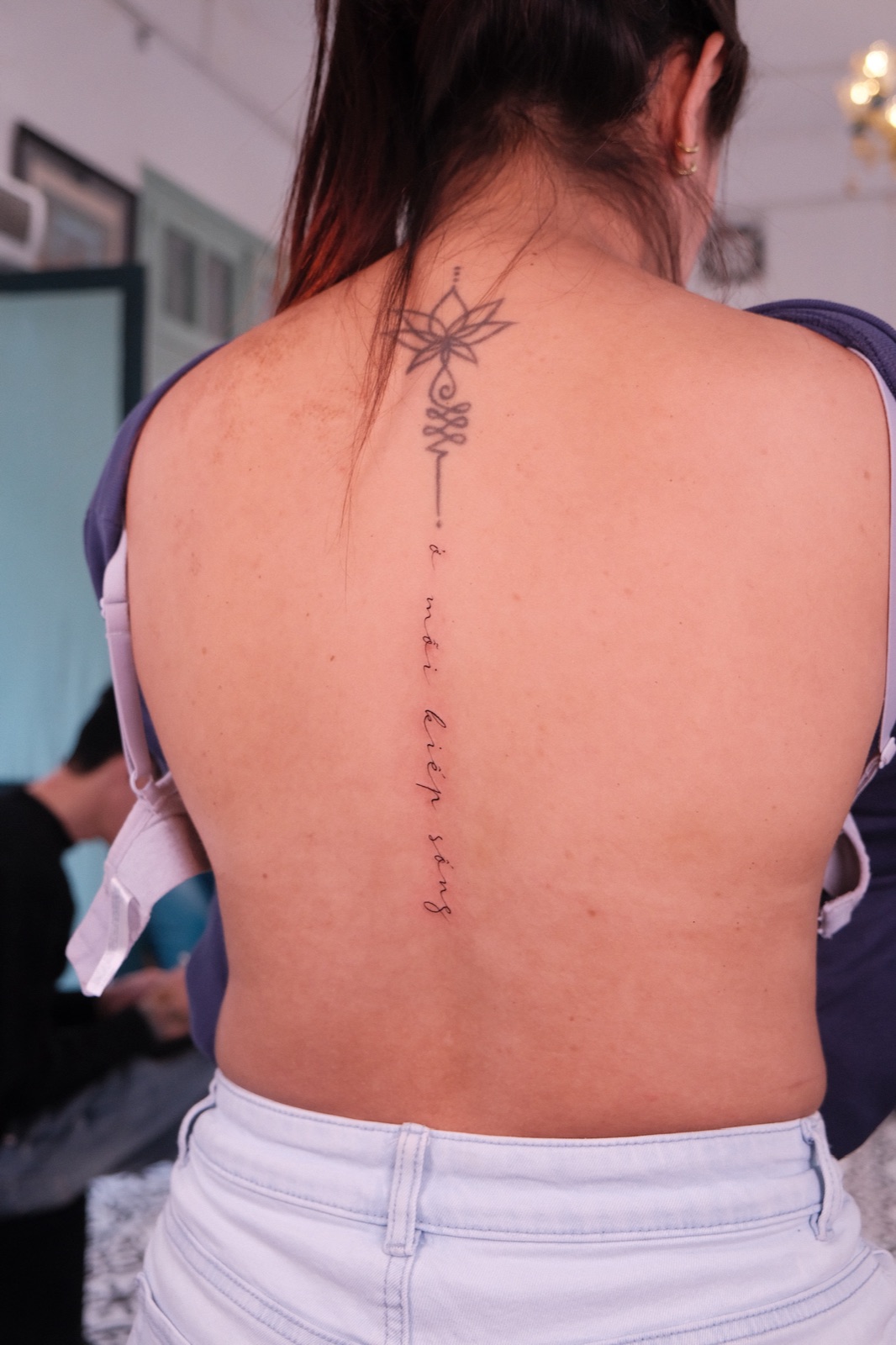 Fine-line lotus flower and Vietnamese script tattoo running down the spine