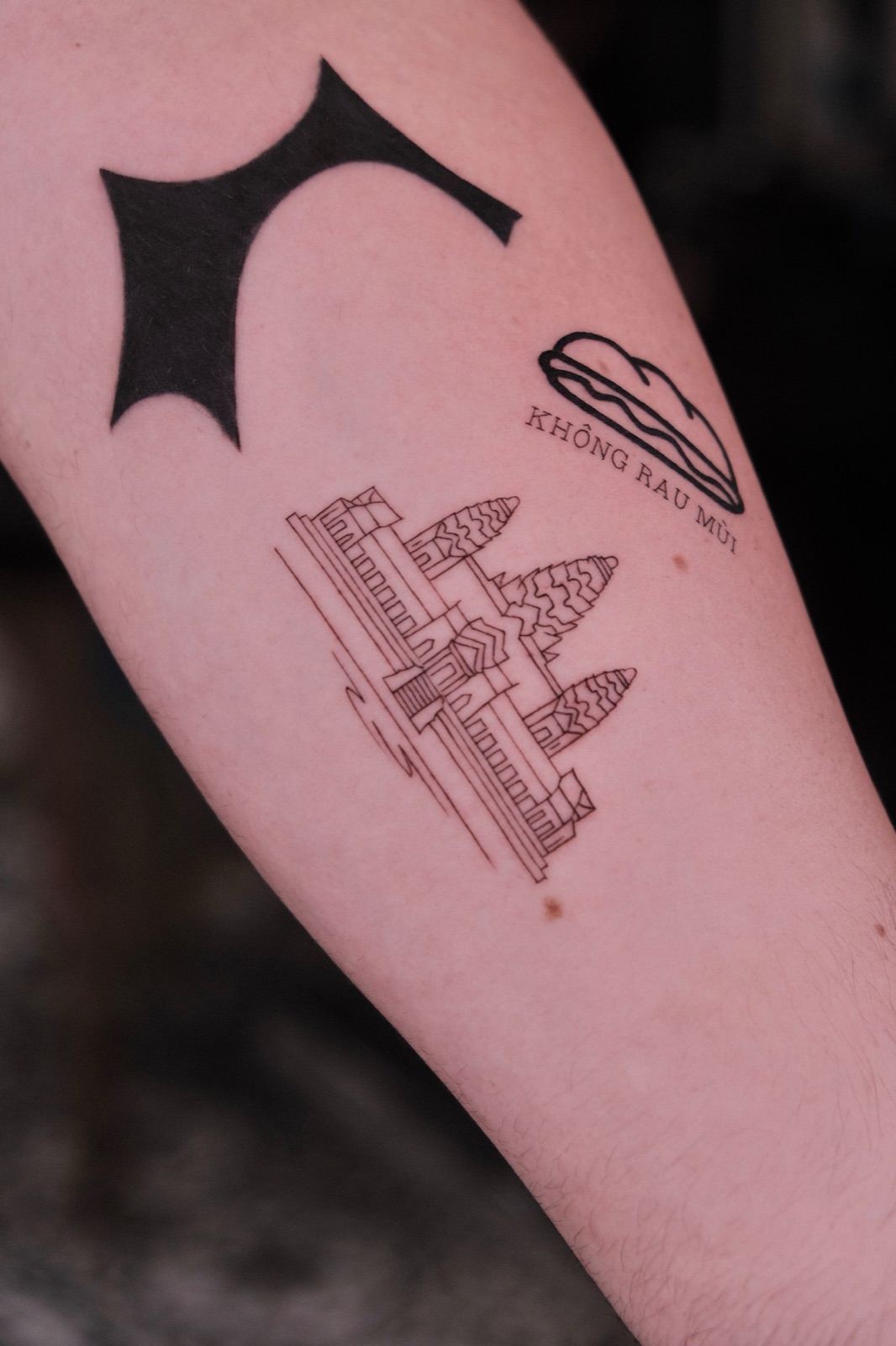 Fine-line tattoo of a market landmark building alongside a no-cilantro banh mi lettering tattoo and a blackwork bat-shaped tattoo on the forearm