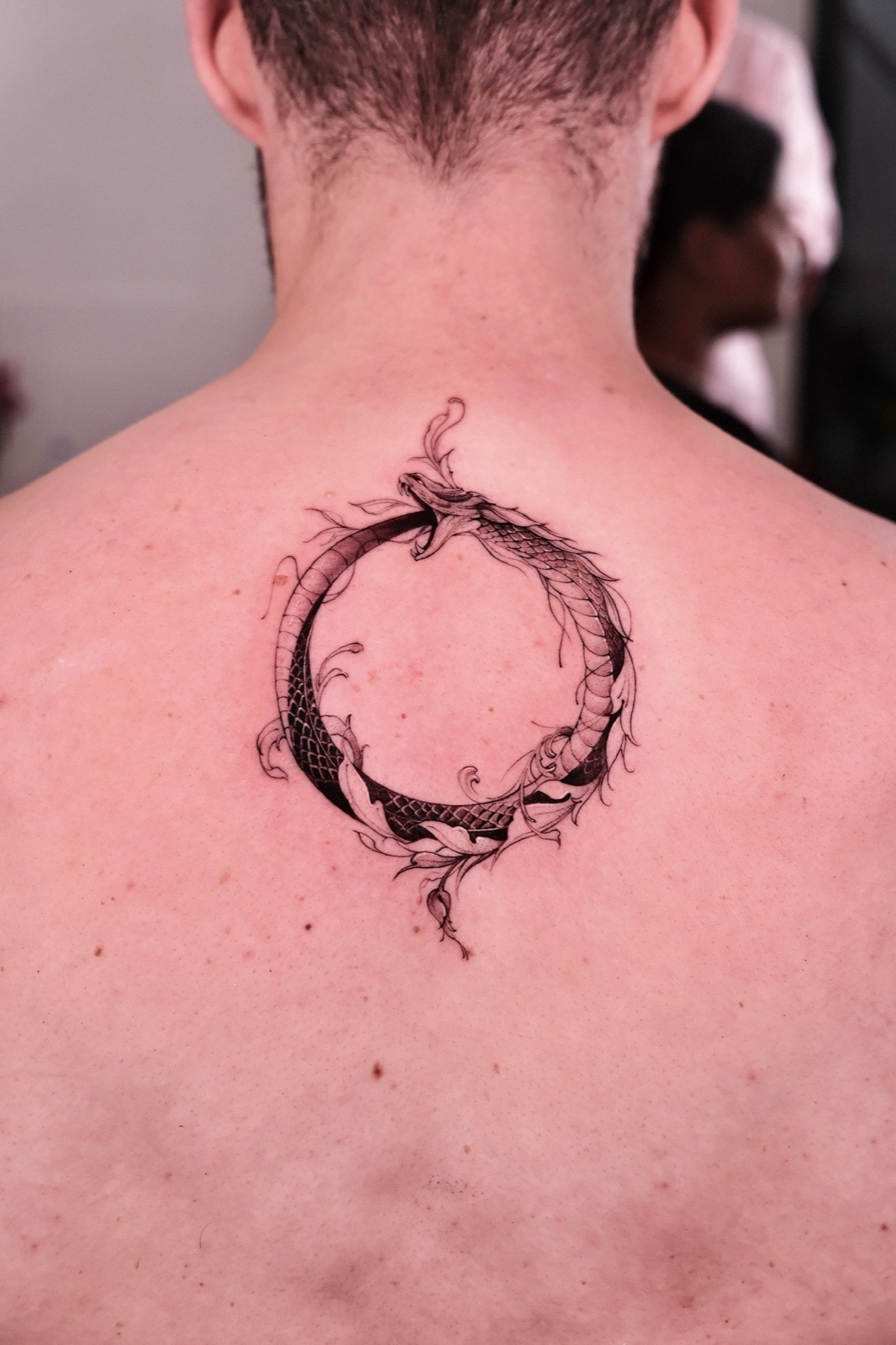 Fine-line ouroboros snake tattoo with leaves on the back of the neck.