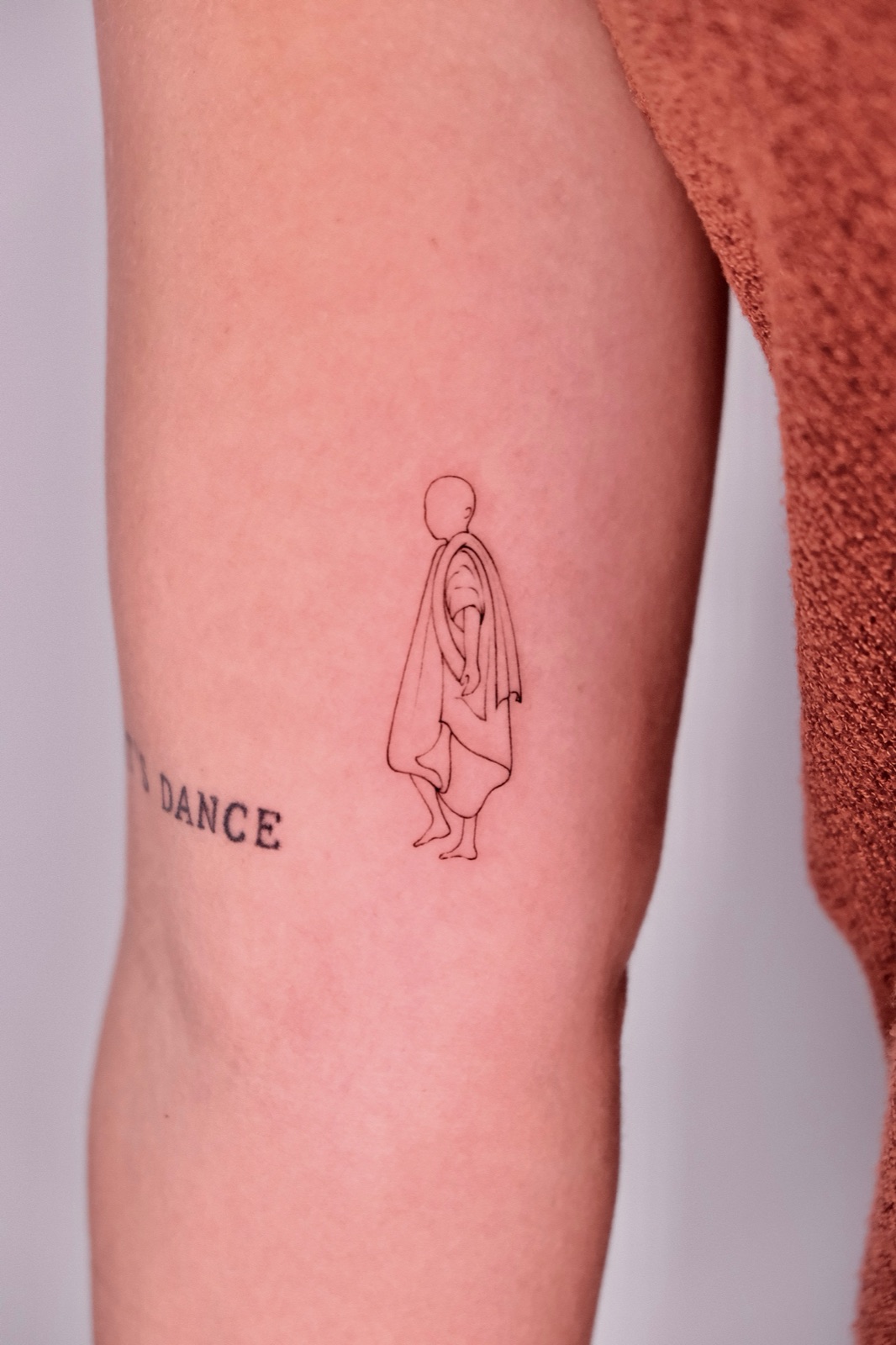 Fine-line silhouette tattoo of a robed figure walking, next to the word DANCE, on the upper arm