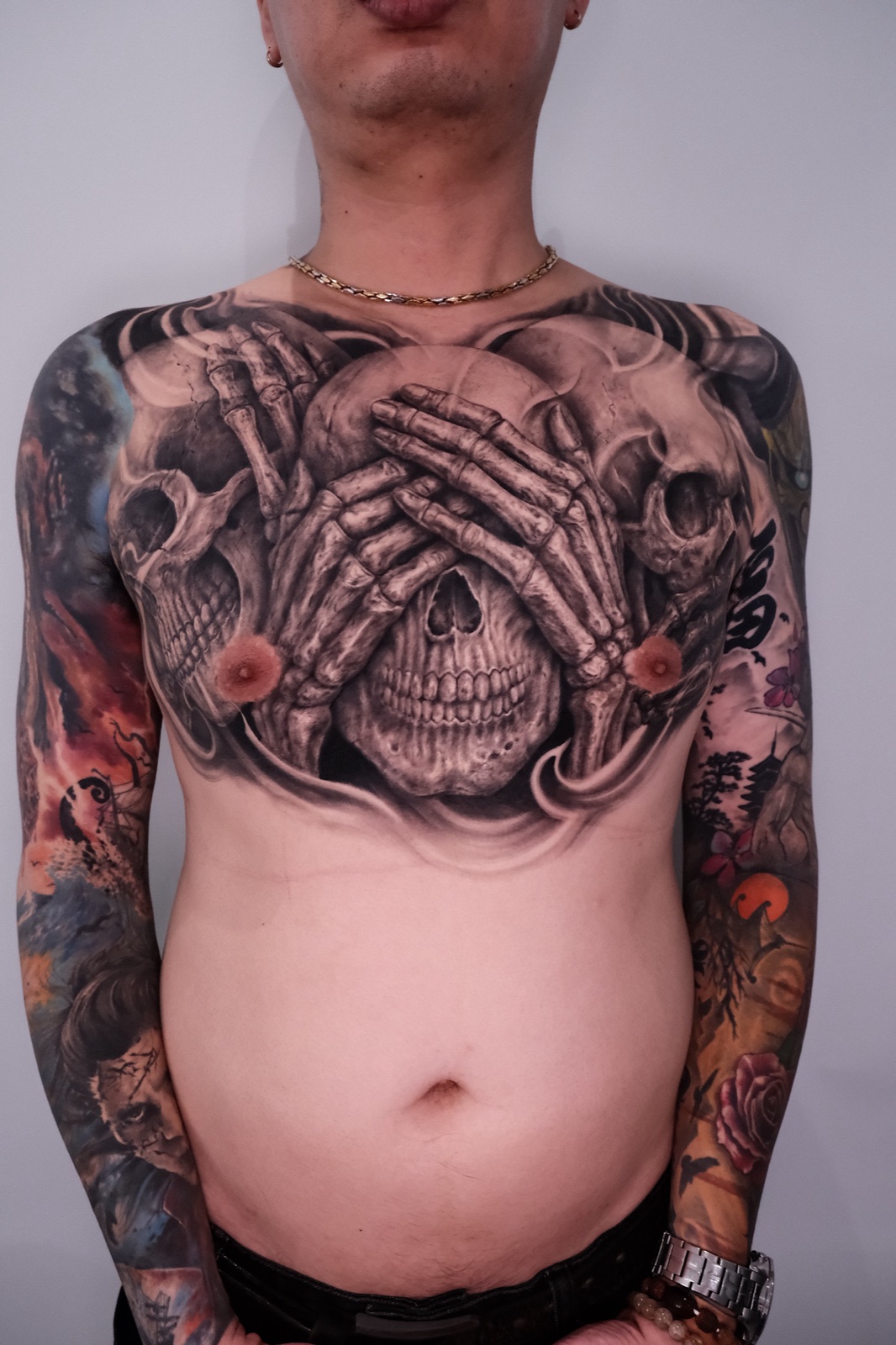 Large black and grey realism chest tattoo of a skull covered by skeletal hands flanked by two more skulls