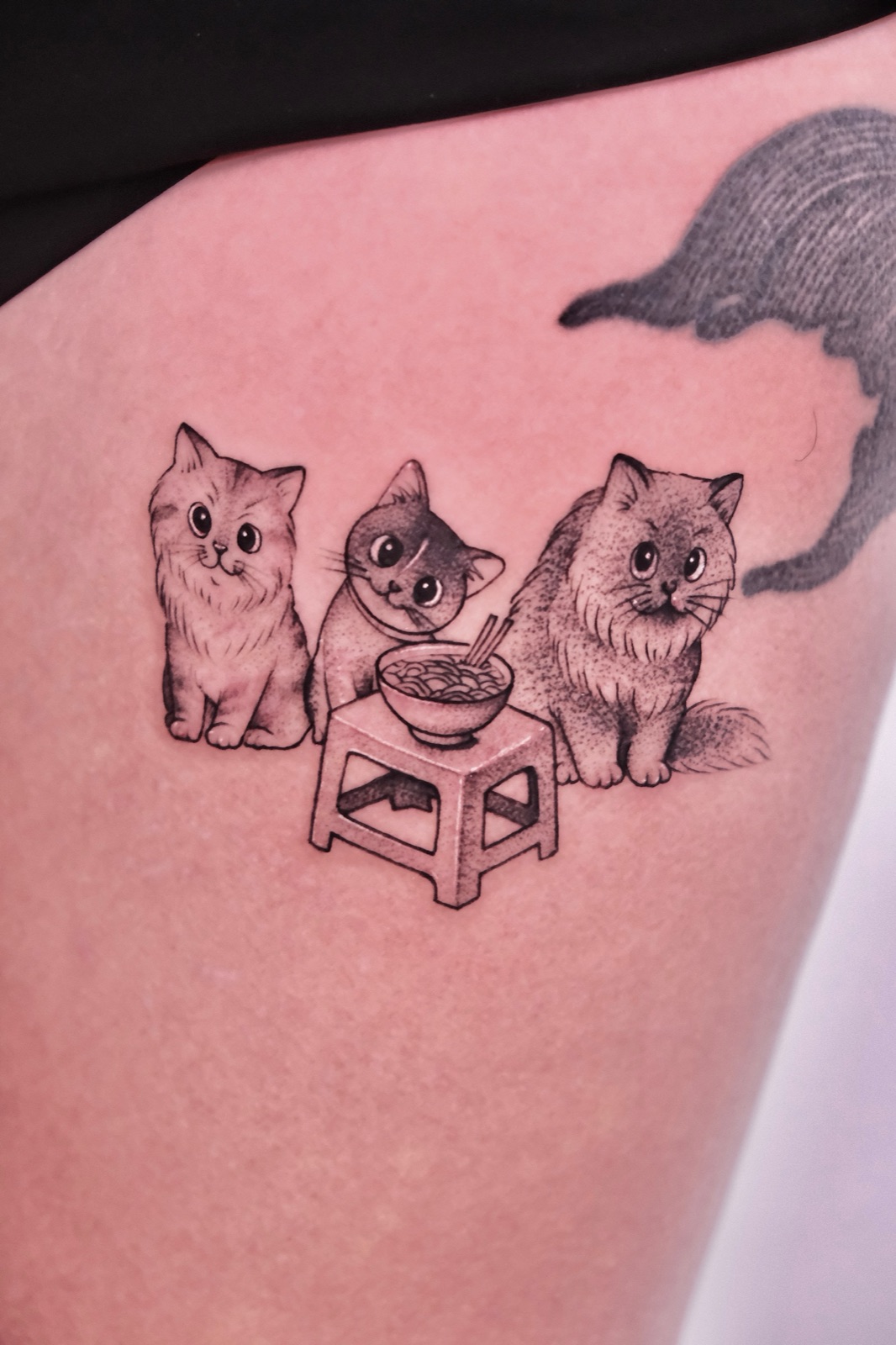 Three cute cartoon cats sitting around a stool with a noodle bowl, black and grey fine tattoo on the thigh near an otter tail tattoo