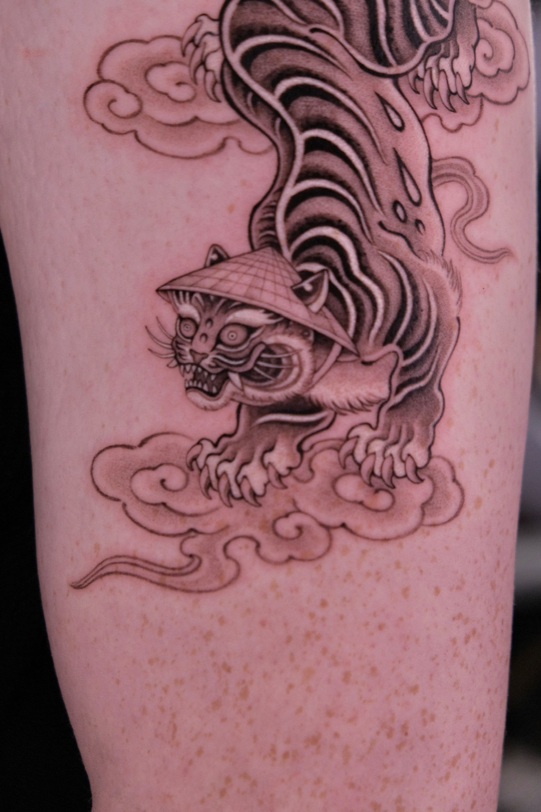 Traditional black-and-grey tiger tattoo wearing a conical hat, crawling through clouds on the upper arm