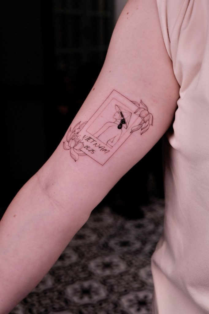 Fine-line polaroid-style tattoo of a woman in a conical hat labeled VIETNAM 2025