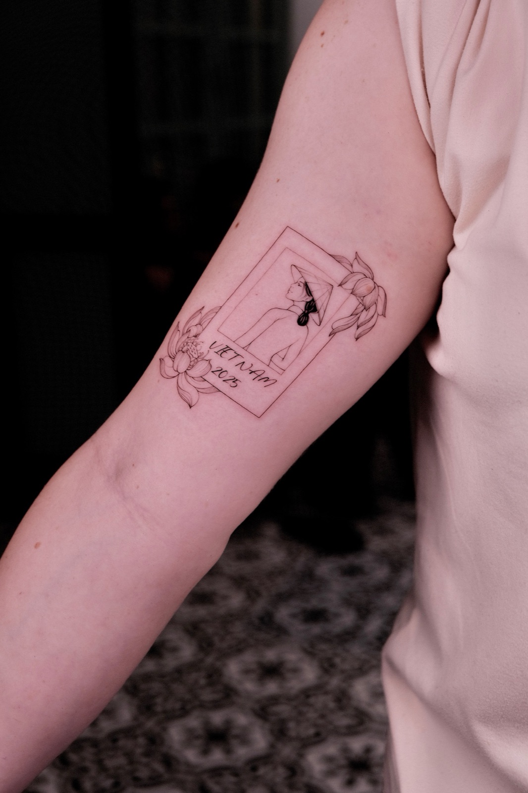 Fine-line polaroid-style tattoo of a woman wearing a conical hat, labeled VIETNAM 2025, tattooed on the upper arm