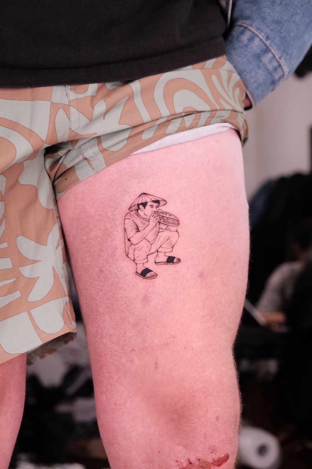 Fine-line illustration tattoo of a Vietnamese man in a conical hat crouching and eating banh mi, on the thigh