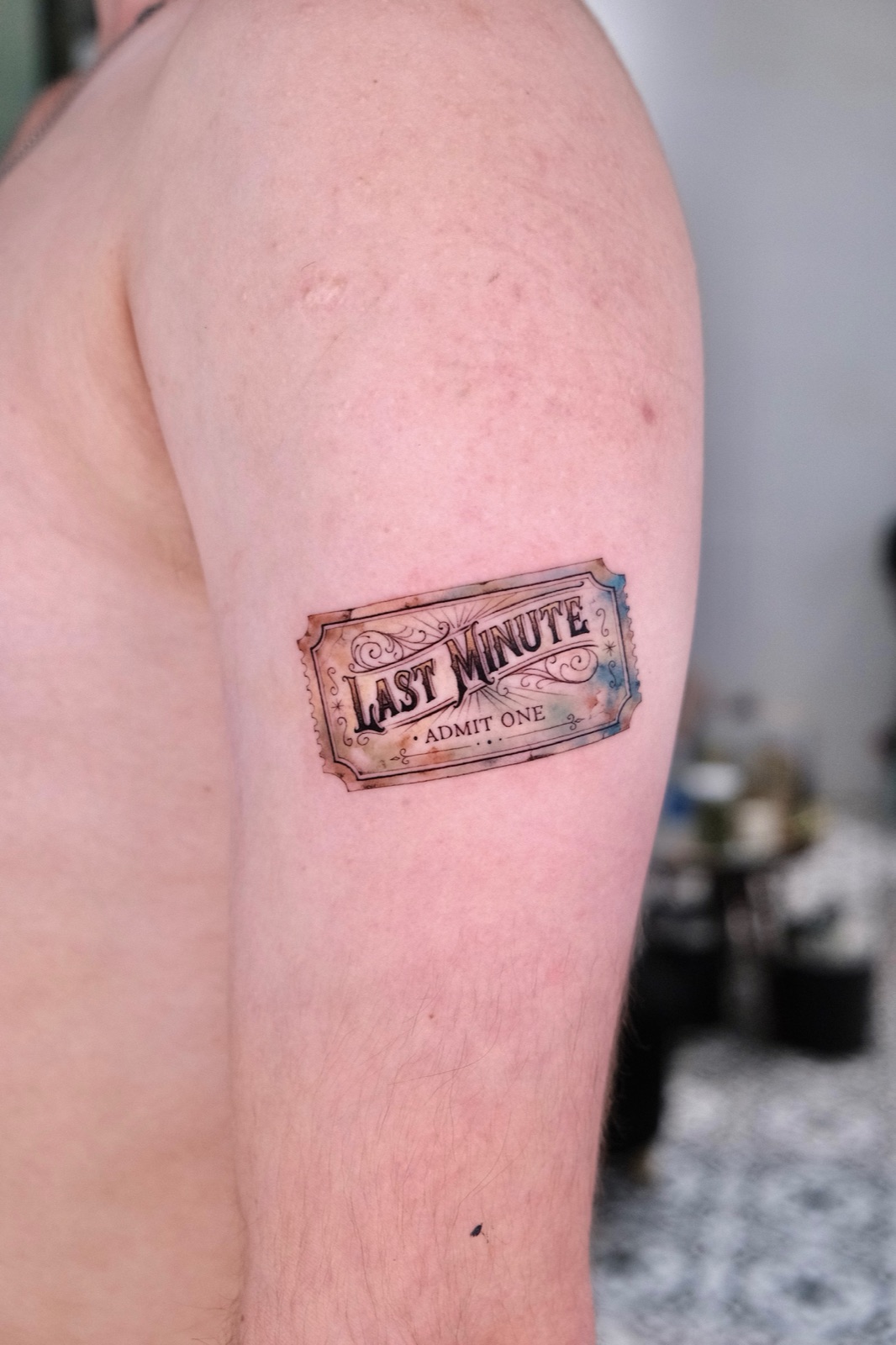 Colorful vintage-style ticket tattoo reading Last Minute Admit One on the upper arm