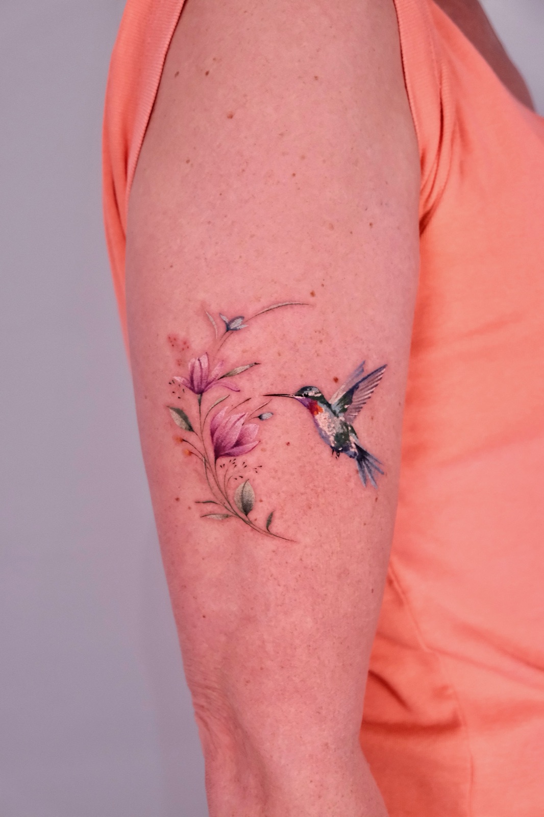 Watercolor-style hummingbird feeding on pink flowers, tattooed on the upper arm