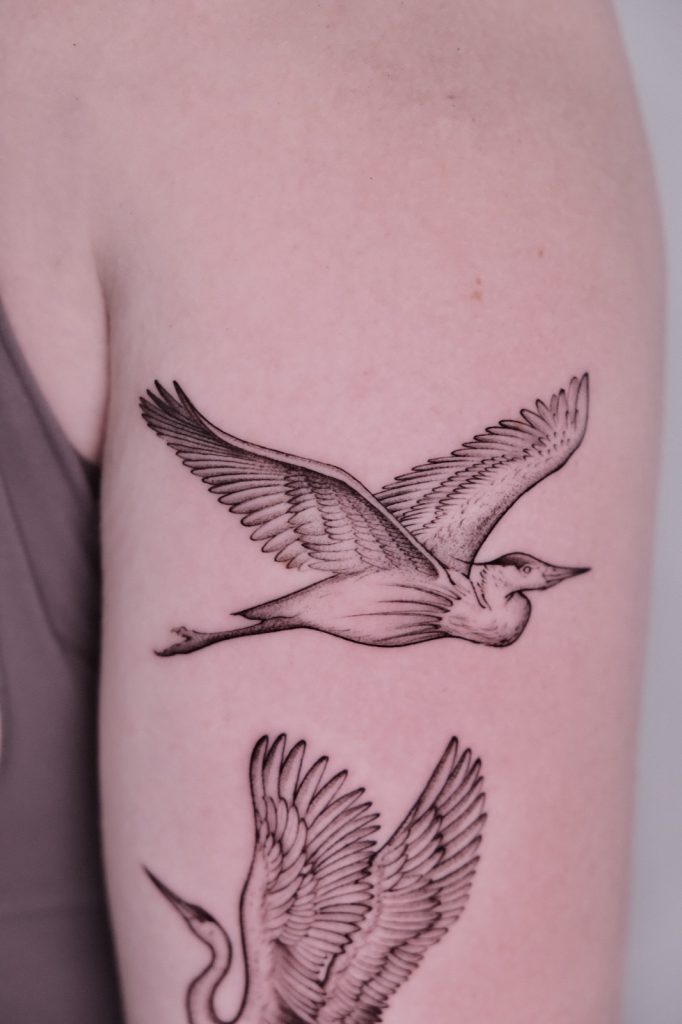 Two fine-line flying cranes tattooed on the upper arm