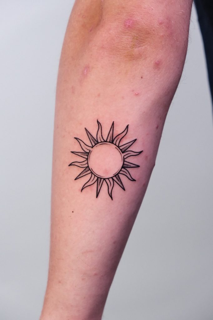 Fine-line sun tattoo with wavy rays on the forearm
