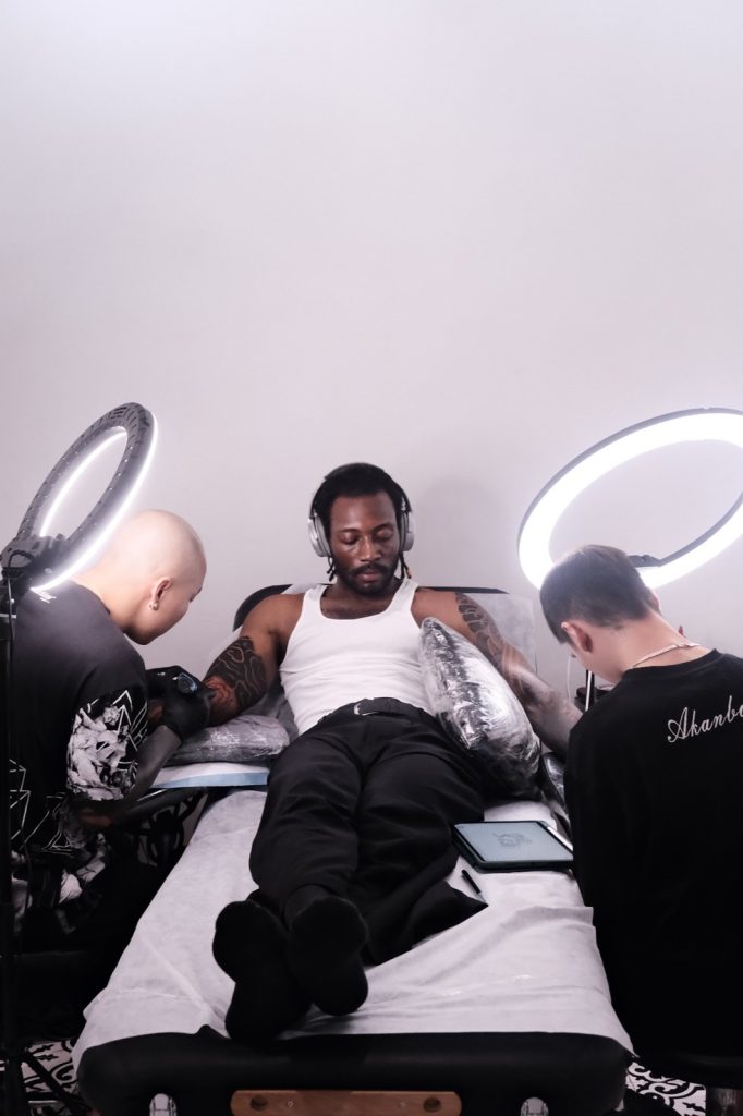 Client in a tank top and loose trousers being tattooed by two artists
