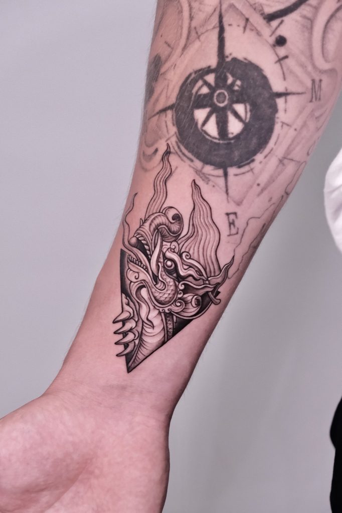 Black and grey compass with a Thai-style guardian head tattooed on the forearm